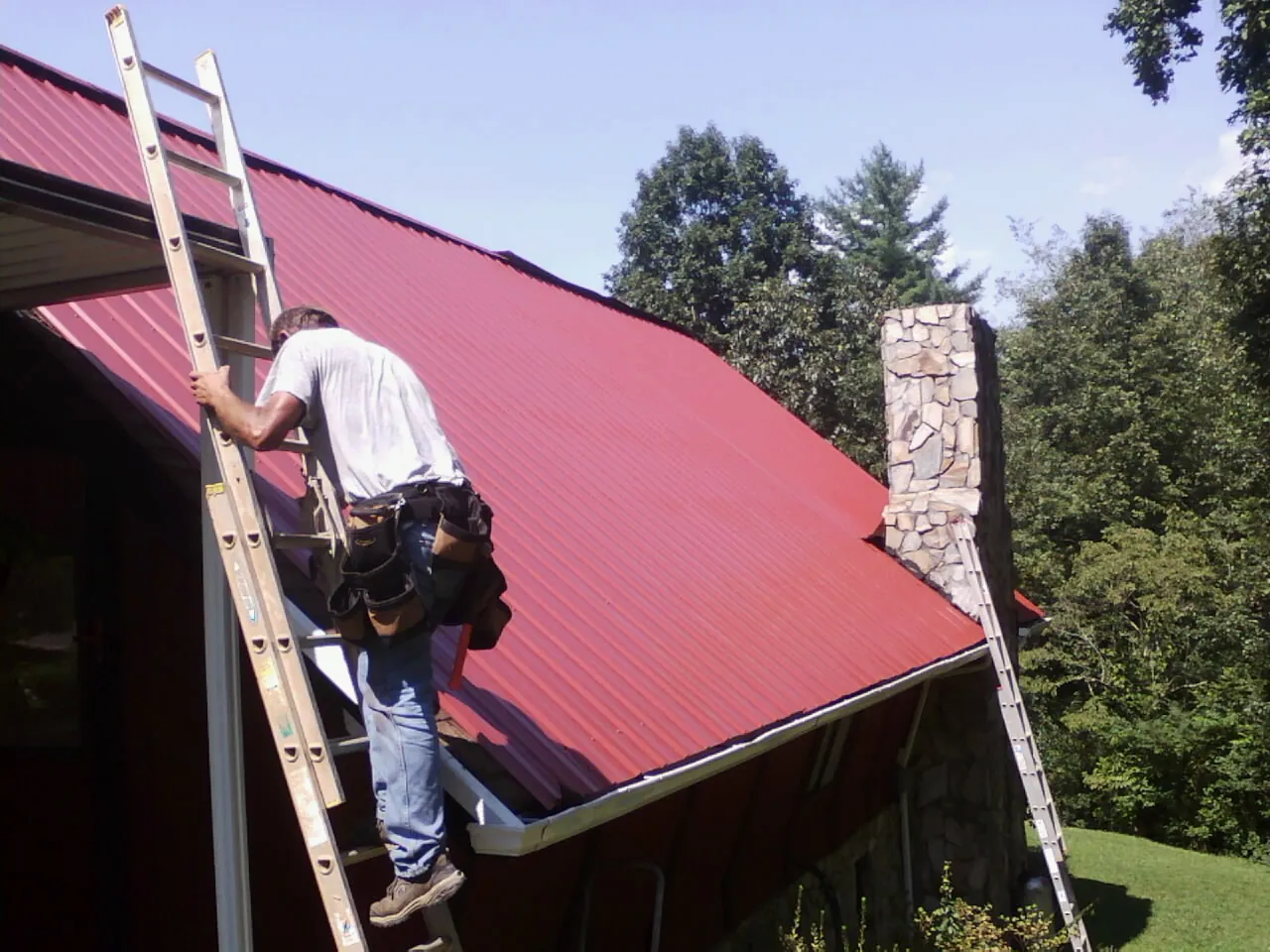 Roofer on a ladder beside a red metal roof and stone chimney — Soffit & Fascia Repair by Roofing Brothers in Conley